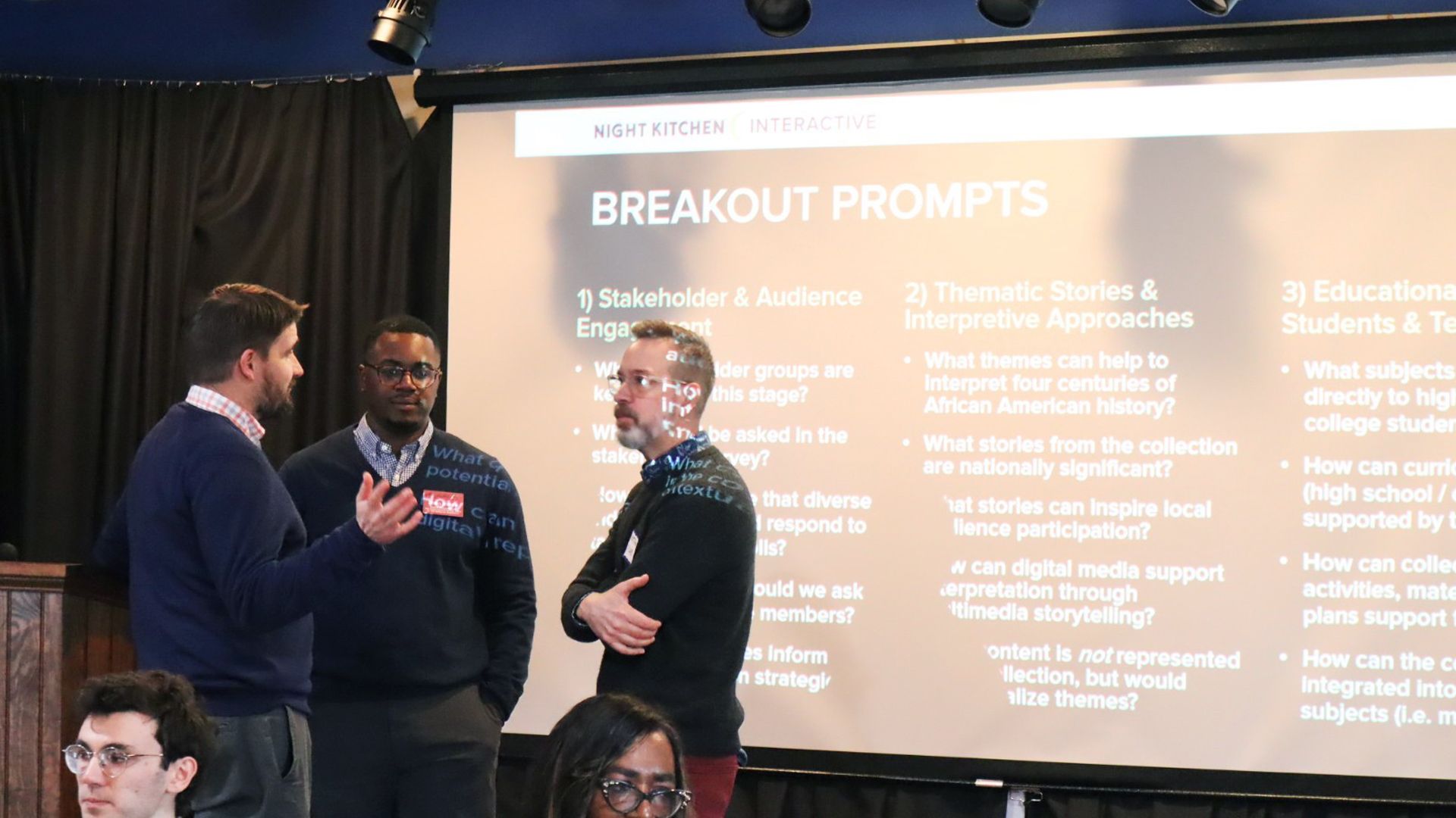 Three men talk in a circle in front of a projector screen that reads "breakout prompts"